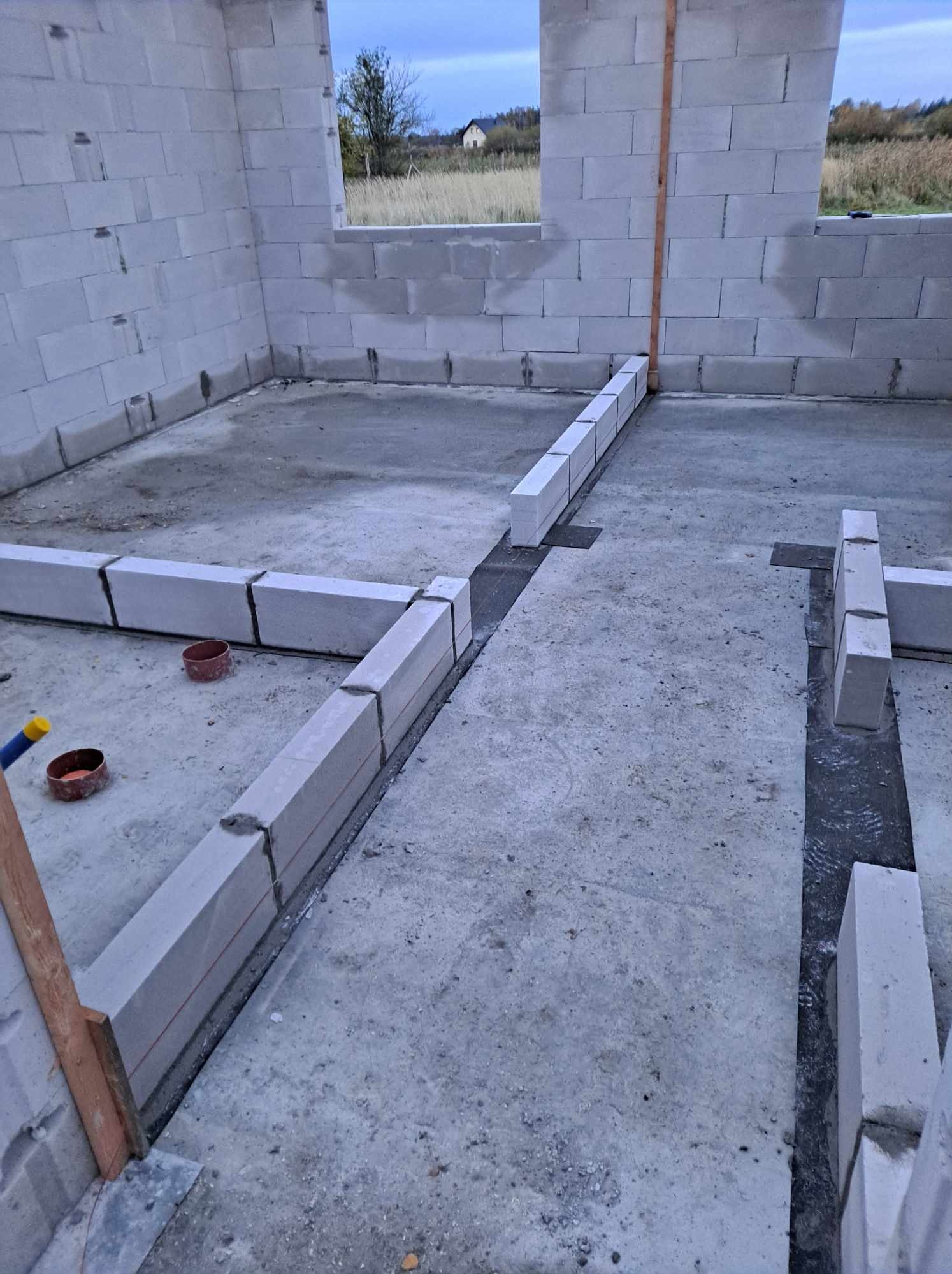 Wnętrze w trakcie budowy z białych bloczków, widoczne fundamenty i ściany w surowym stanie, dwa czerwone pojemniki na podłodze.