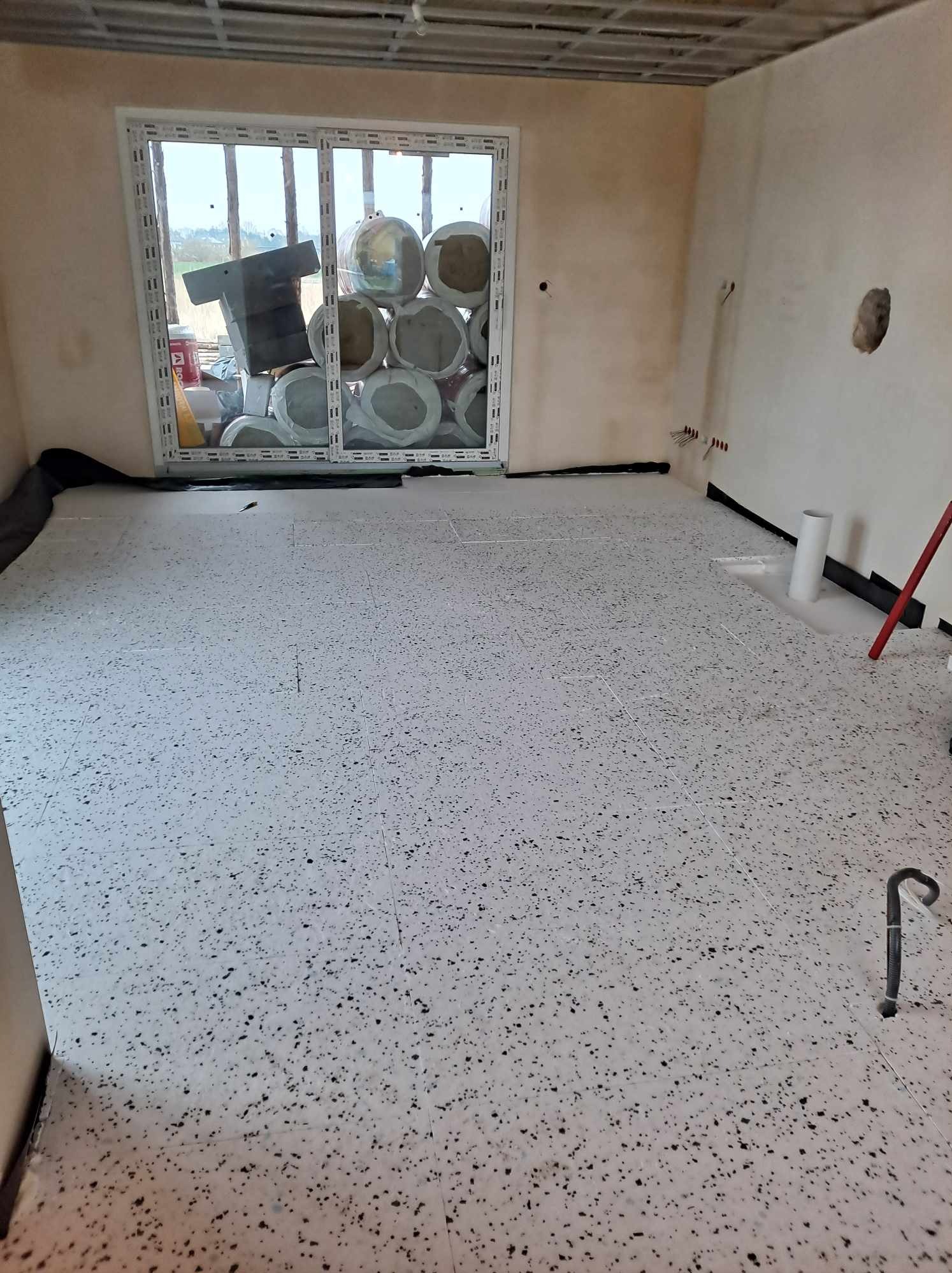 Przygotowane pomieszczenie do dalszych prac wykończeniowych: podłoga pokryta płytami izolacyjnymi w czarno-białe kropki, okno z widocznymi materiałami budowlanymi i izolacyjnymi, ściany...