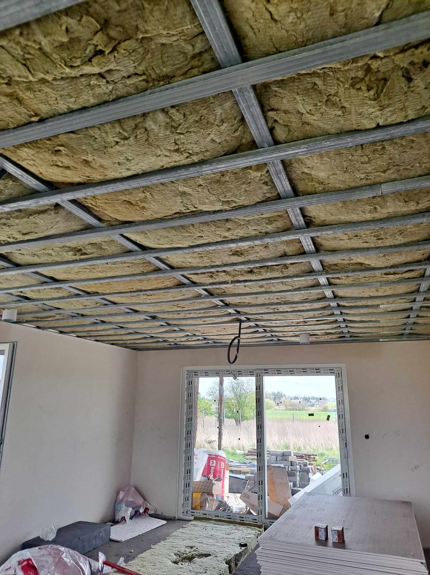 Montaż izolacji z wełny mineralnej w metalowym stelażu sufitu podwieszanego, widok z dołu na konstrukcję z widocznymi elementami instalacji elektrycznej i stertą płyt gipsowo-kartonowych w tle.