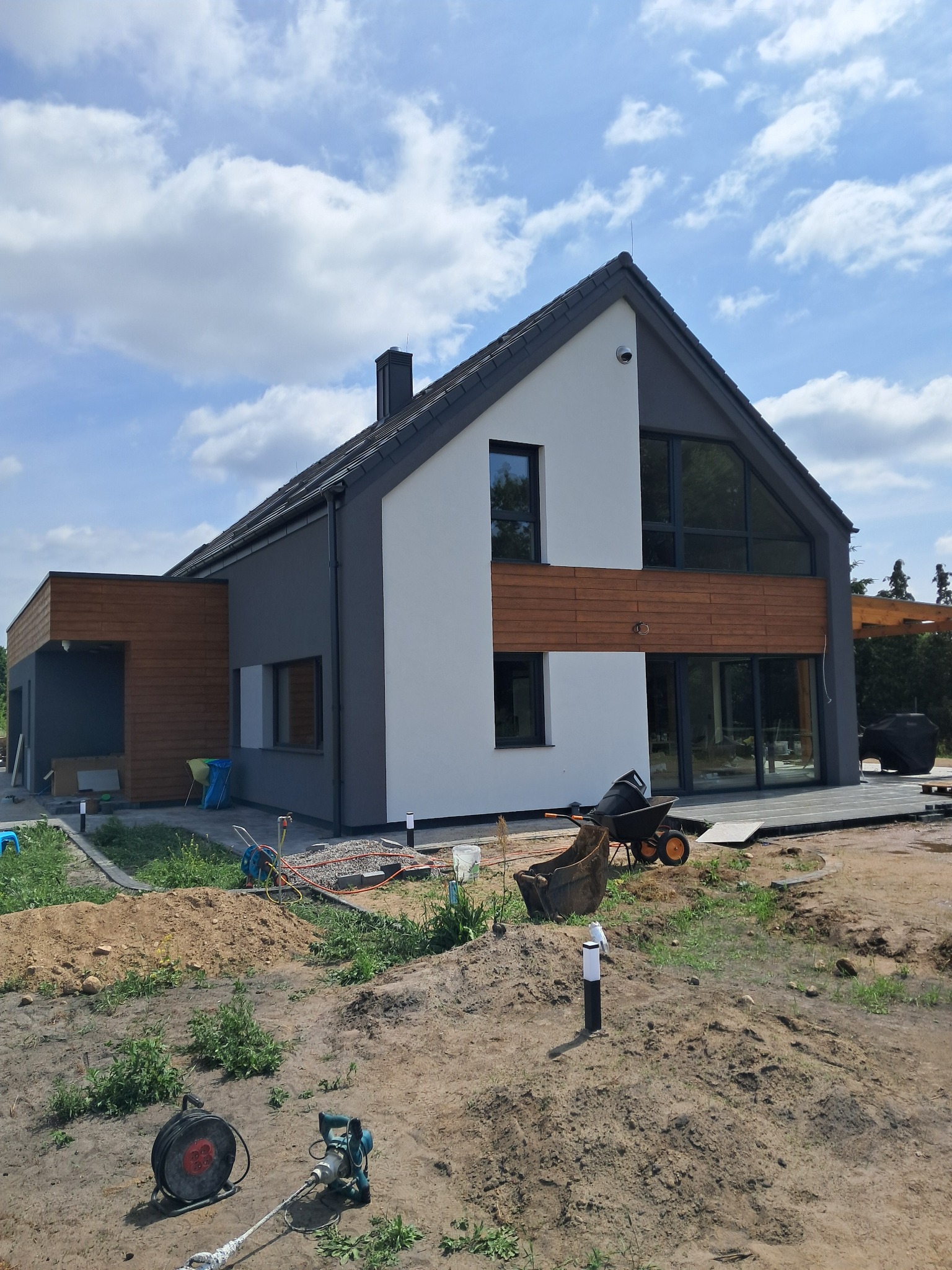 Nowoczesny dom z elewacją w odcieniach szarości i bieli, z drewnianymi akcentami. Wokół budynku widoczne narzędzia i materiały budowlane.