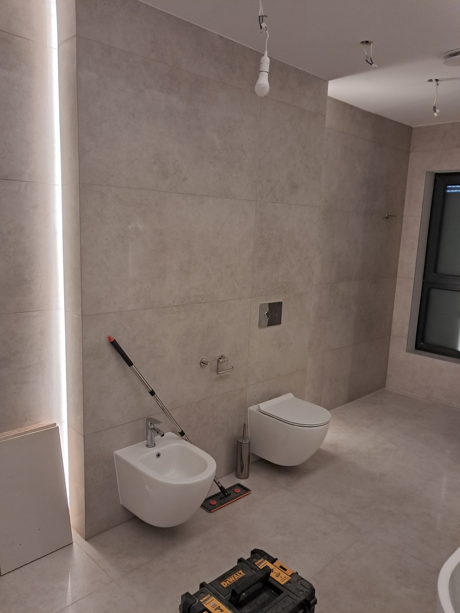 Wykończona łazienka z beżowymi płytkami, wiszącą toaletą i bidetem. Widoczna skrzynka narzędziowa DeWalt, sugerująca prace wykończeniowe w toku. Minimalistyczny design.