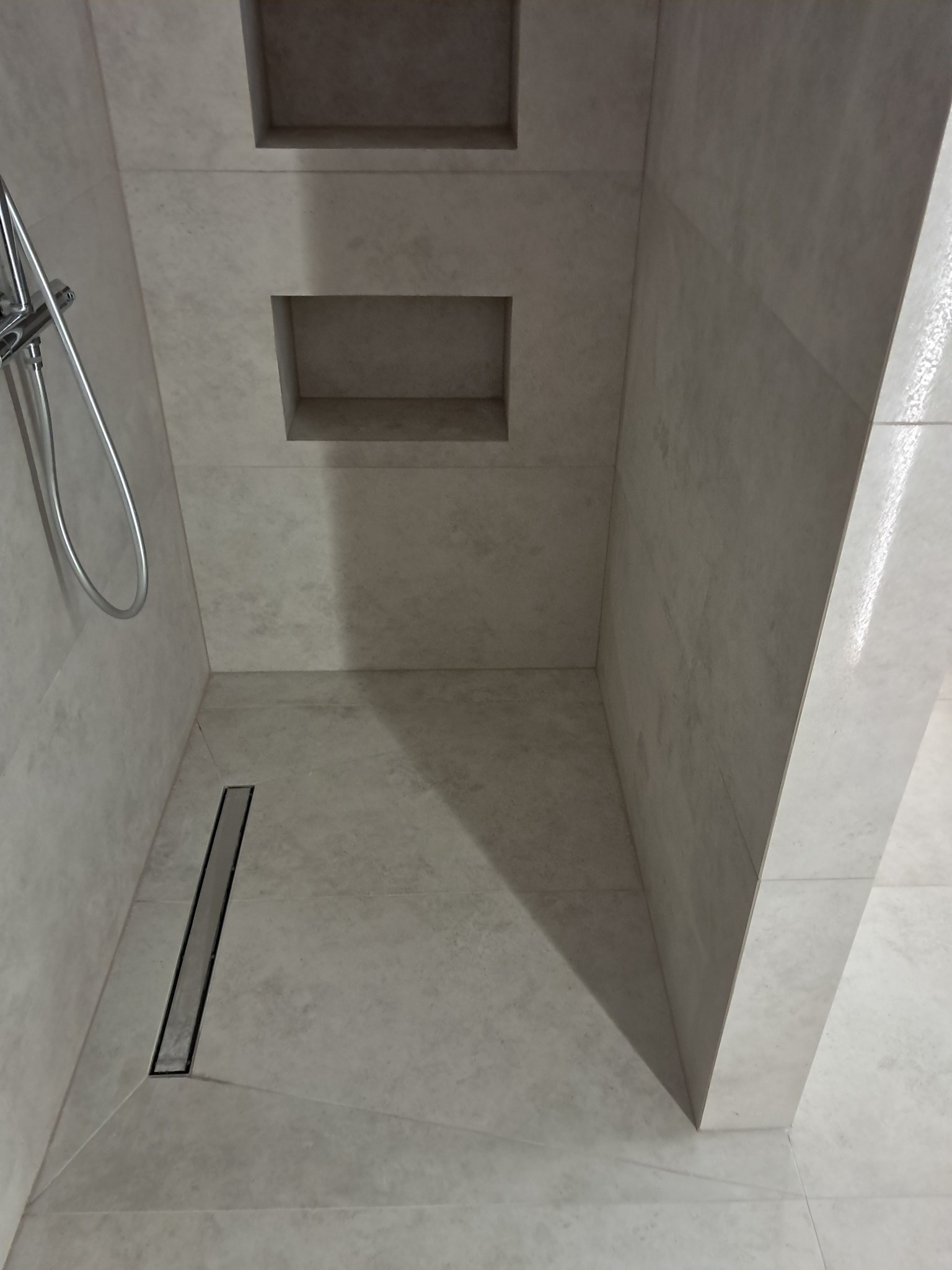 Wykończona kabina prysznicowa z odpływem liniowym i półkami w ścianie, wyłożona jasnymi płytkami. Widoczny fragment słuchawki prysznicowej.