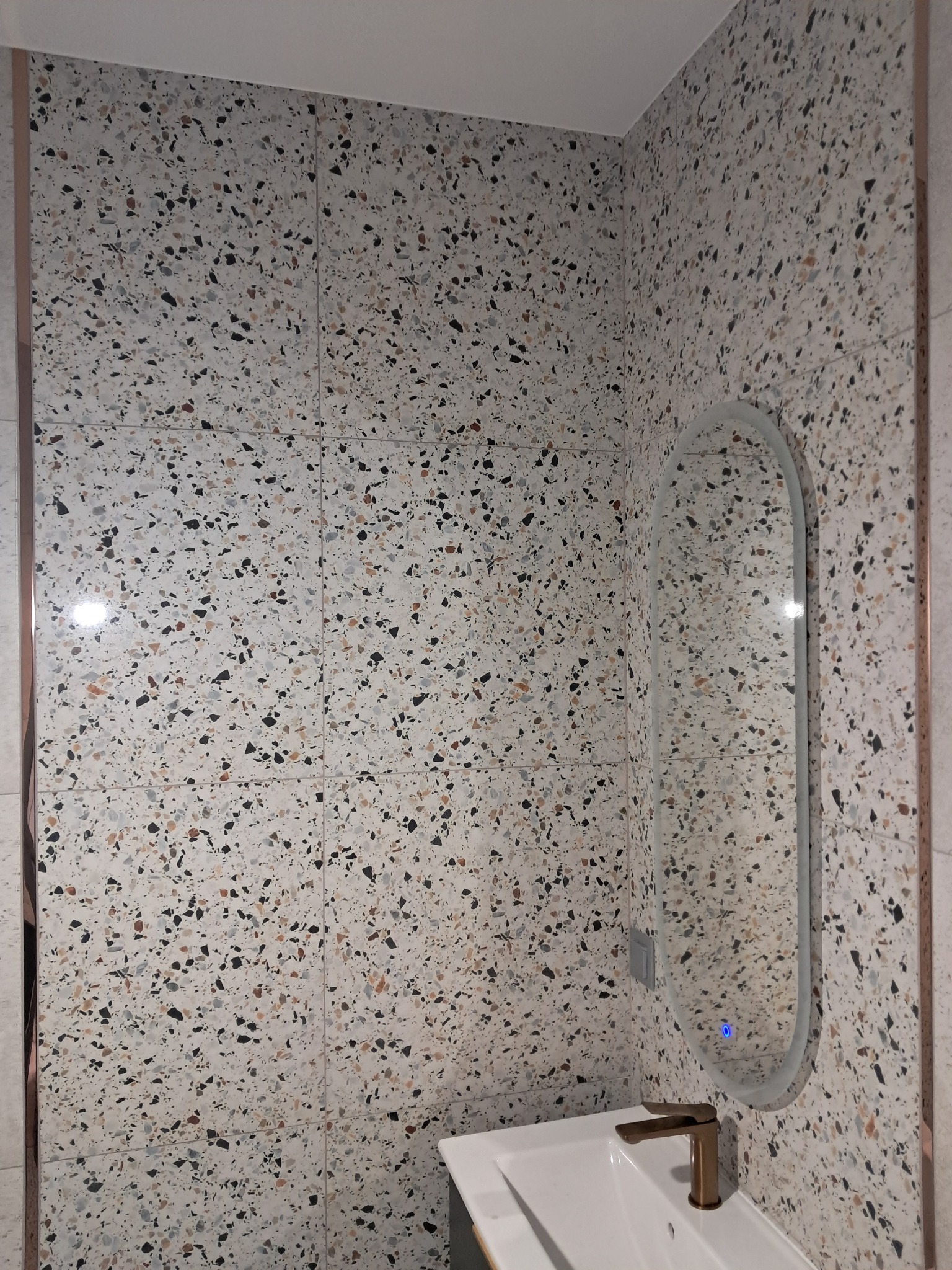 Wykończona ściana łazienki z terazzo, z owalnym lustrem podświetlanym led i umywalką z baterią w kolorze miedzi. Widoczne fugi i detale wykończenia.