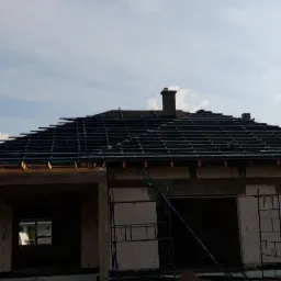 Budowa domu w stanie surowym otwartym: widoczna konstrukcja dachu z łatami, ściany z białego bloczka, rusztowania przy oknach, betonowy szkielet werandy, komin. W tle drzewa i błękitne niebo...