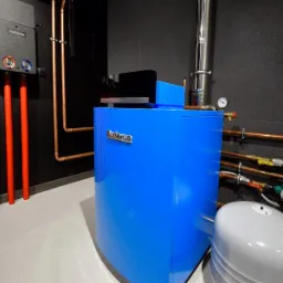 Niebieski kocioł grzewczy Buderus z podłączonymi rurami miedzianymi i czerwonymi, na tle czarnej ściany z szarą podłogą.