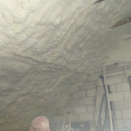 CLEAN-PUR Paweł Mateusiak - Poddasze po nałożeniu izolacji pianką poliuretanową, widoczne nierówności i struktura piany, fragment drewnianej konstrukcji dachu oraz pracownik w czapce w tle.