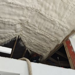 CLEAN-PUR Paweł Mateusiak - Poddasze z widoczną strukturą natryśniętej pianki izolacyjnej, mocowanej do krokwi za pomocą szpilek. W tle widoczny pracownik w trakcie aplikacji. Fragment drabiny i ściany w dolnej części kadru.