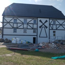 CLEAN-PUR Paweł Mateusiak - Budynek po renowacji elewacji z charakterystycznym wzorem szachownicy i wiatraka, widoczne materiały budowlane i rusztowanie w tle.