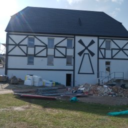 CLEAN-PUR Paweł Mateusiak - Budynek z białą elewacją i czarnym szkieletem konstrukcyjnym, ozdobiony motywem wiatraka, w trakcie prac remontowych z widocznymi materiałami izolacyjnymi i gruzem.