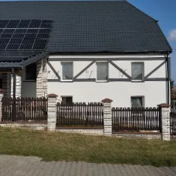 Dom jednorodzinny z panelami słonecznymi na dachu i elewacją w stylu muru pruskiego, okna zabezpieczone folią malarską, ogrodzenie drewniano-murowane.