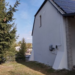 CLEAN-PUR Paweł Mateusiak - Boczny widok otynkowanego domu z panelami słonecznymi na dachu, widoczne elementy instalacji elektrycznej na elewacji, obok domu rosną drzewa iglaste.
