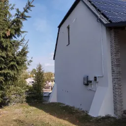 Boczny widok otynkowanego domu z panelami słonecznymi na dachu, widoczne elementy instalacji elektrycznej na elewacji, obok domu rosną drzewa iglaste.