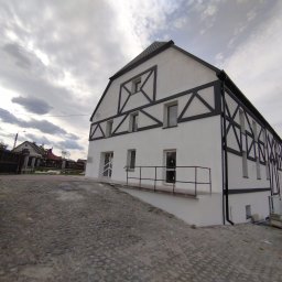 CLEAN-PUR Paweł Mateusiak - Odnowiony budynek z białą elewacją ozdobioną czarnymi, geometrycznymi elementami dekoracyjnymi, widoczny z poziomu brukowanej posesji w pochmurny dzień.