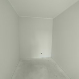 CLEAN-PUR Paweł Mateusiak - Pusty, świeżo pomalowany pokój z szarymi ścianami i betonową podłogą, widoczne gniazdko elektryczne i listwa przypodłogowa, surowy stan wykończenia.