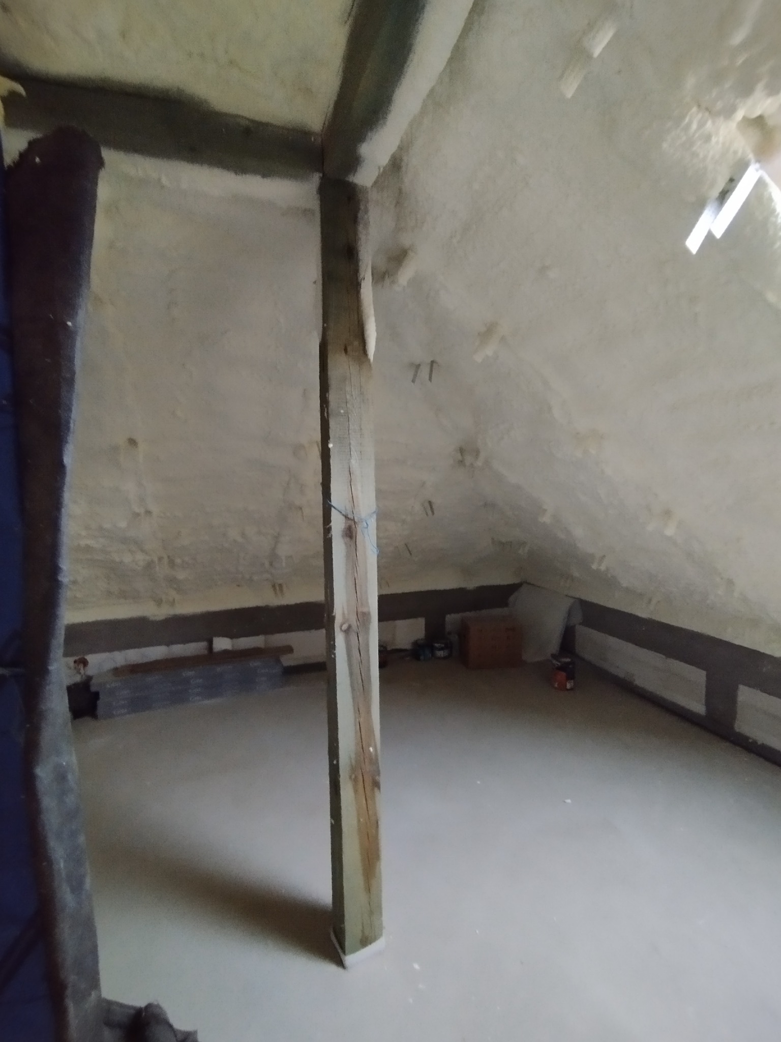 Poddasze po natrysku pianki izolacyjnej, widoczna drewniana konstrukcja i fragment podłogi, z elementami wyposażenia w tle.