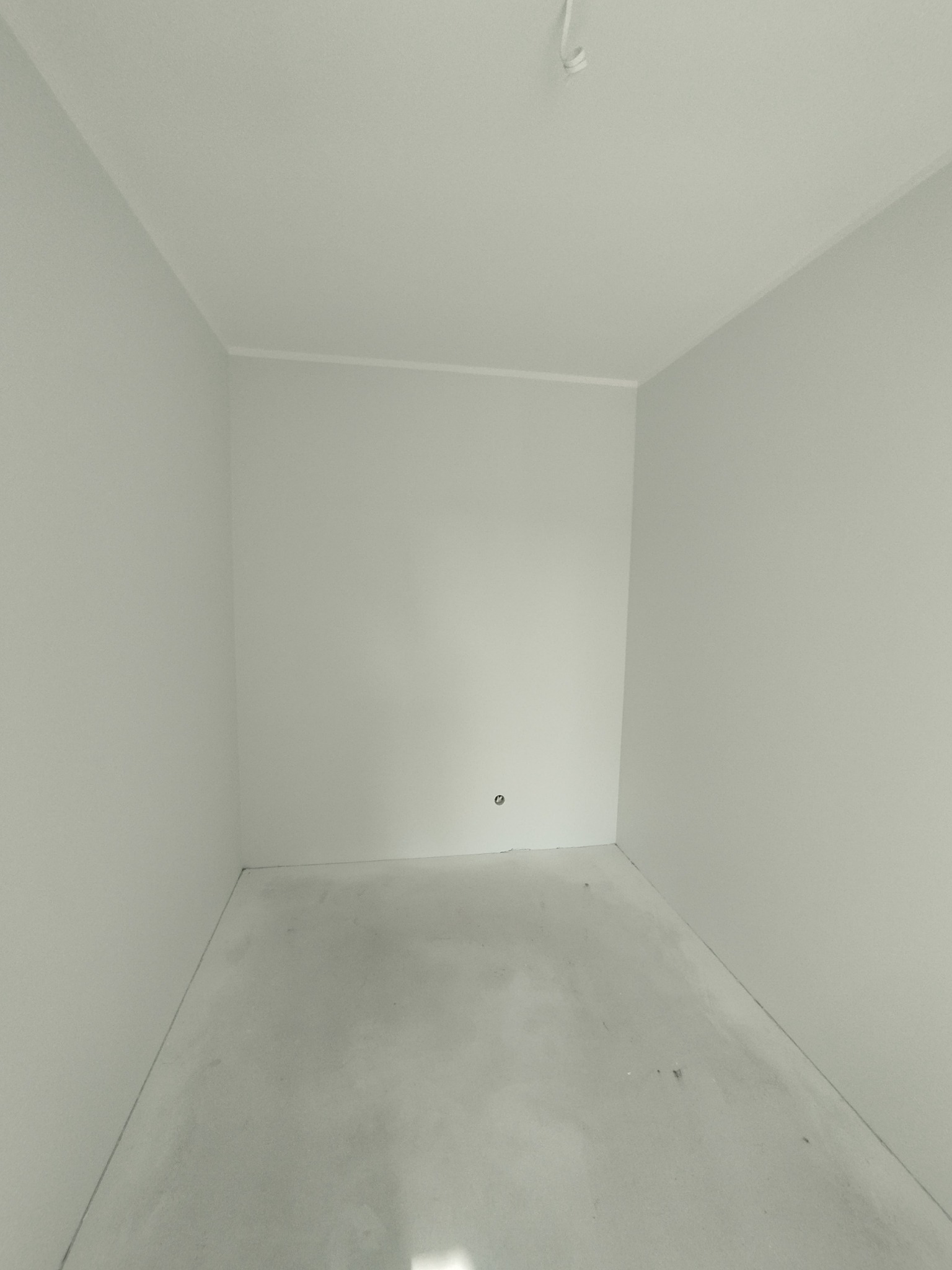 Pusty, świeżo pomalowany pokój z szarymi ścianami i betonową podłogą, widoczne gniazdko elektryczne i listwa przypodłogowa, surowy stan wykończenia.