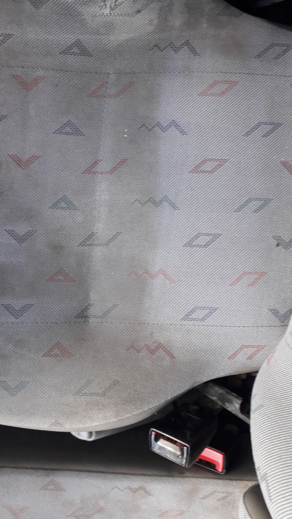 Fragment tapicerki samochodowej przed i po czyszczeniu, widoczna różnica w jasności i usunięciu zabrudzeń, wzór geometryczny z trójkątów, zygzaków i rombów w kolorach czerwonym, niebieskim...