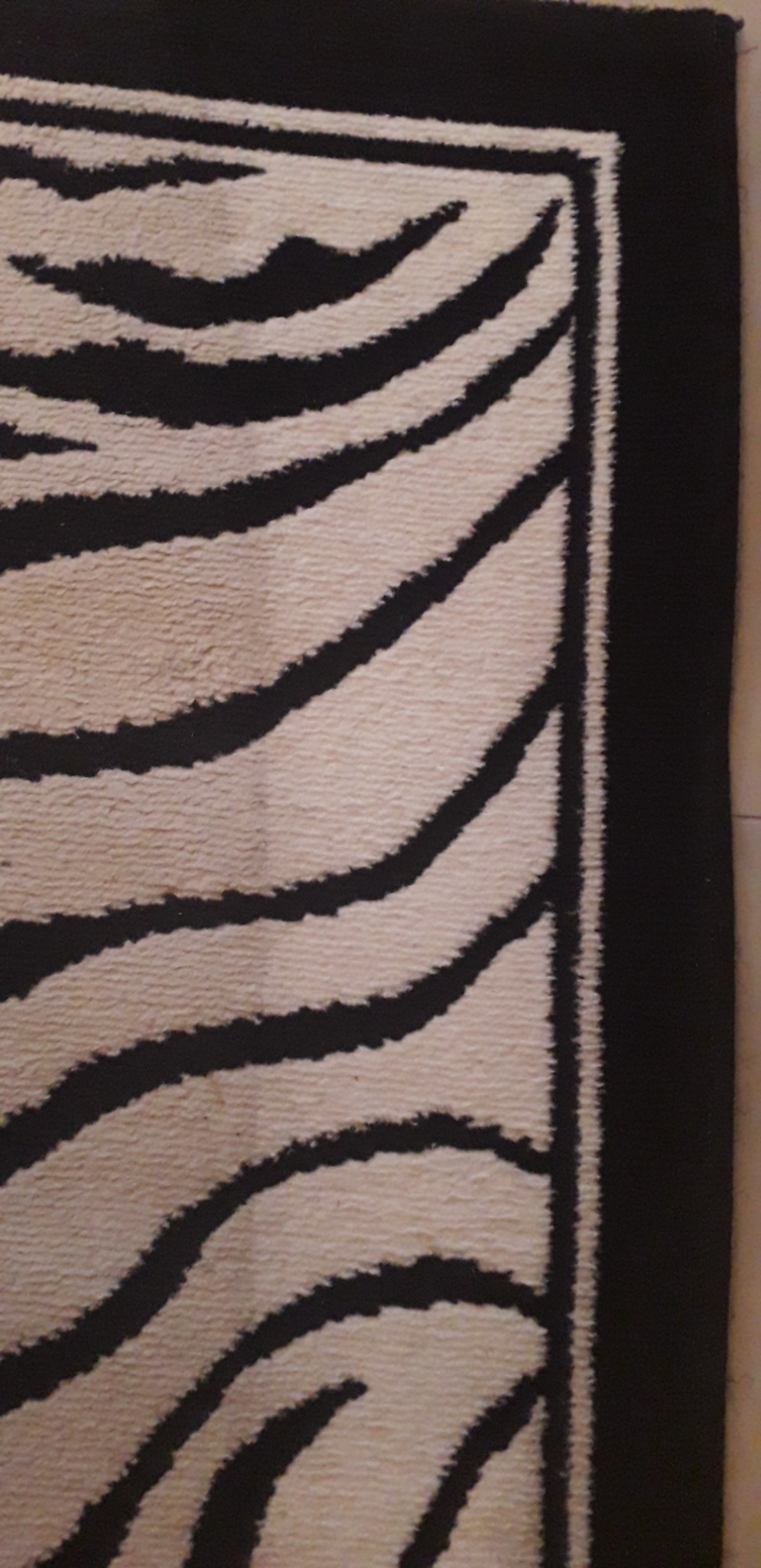 Fragment kremowego dywanu z czarnym, abstrakcyjnym wzorem przypominającym pasy zebry, obramowanego szeroką, czarną bordiurą.