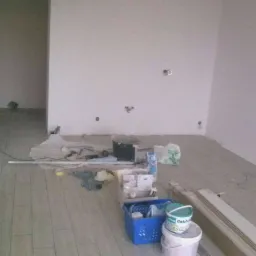 Pokój w trakcie prac wykończeniowych: położona podłoga z paneli, ściany przygotowane do malowania, widoczne narzędzia i materiały budowlane, w tym puszki z farbą 'Dekoral' i niebieski pojemnik...