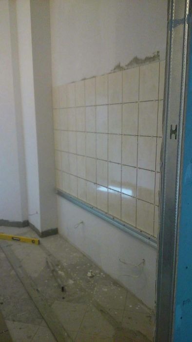 Ściana w trakcie kładzenia płytek ceramicznych w kuchni, widoczne profile metalowe, poziomica leżąca na podłodze i wystające kable elektryczne.