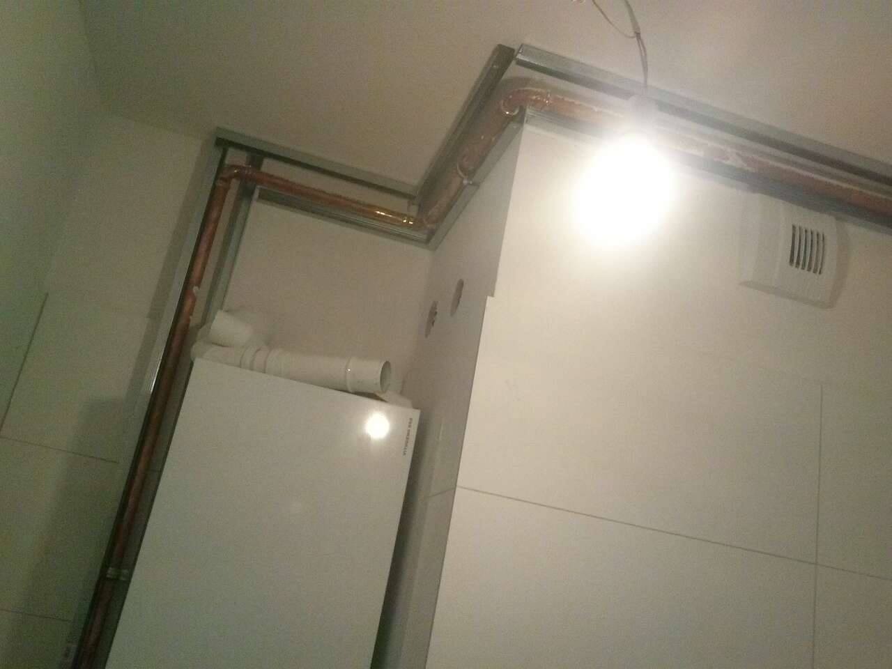 Biały piec grzewczy z podłączeniami rur miedzianych i elementów wentylacyjnych, zamontowany w rogu pomieszczenia wykończonego białymi płytkami, widoczne elementy konstrukcji sufitu podwieszanego.