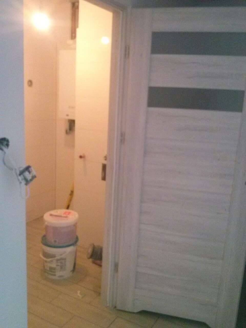 Otwarta, biała, wewnętrzna stolarka drzwiowa z poziomymi, szarymi pasami, widok na fragment łazienki z wiadrami farby na podłodze z płytek.