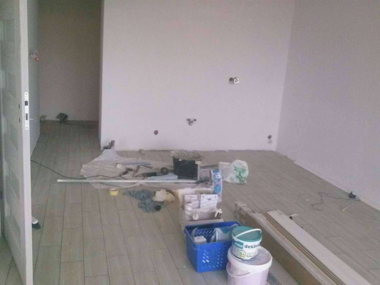 Pokój w trakcie prac wykończeniowych: położona podłoga z paneli, ściany przygotowane do malowania, widoczne narzędzia i materiały budowlane, w tym puszki z farbą 'Dekoral' i niebieski pojemnik...