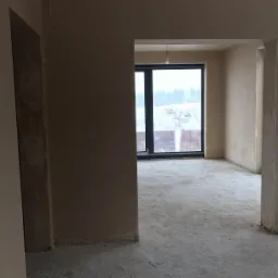 Surowe wnętrze po nałożeniu tynków maszynowych, widoczne ślady niedoskonałości i wykończenia przy podłodze, z oknem na zimowy krajobraz w tle.