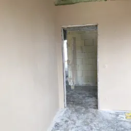 Wnętrze w trakcie prac wykończeniowych: ściany pokryte świeżo nałożoną, jasną warstwą, widoczne surowe drewno stropu oraz fragment ściany z pustaków przez otwór drzwiowy, podłoga z resztkami zaprawy.