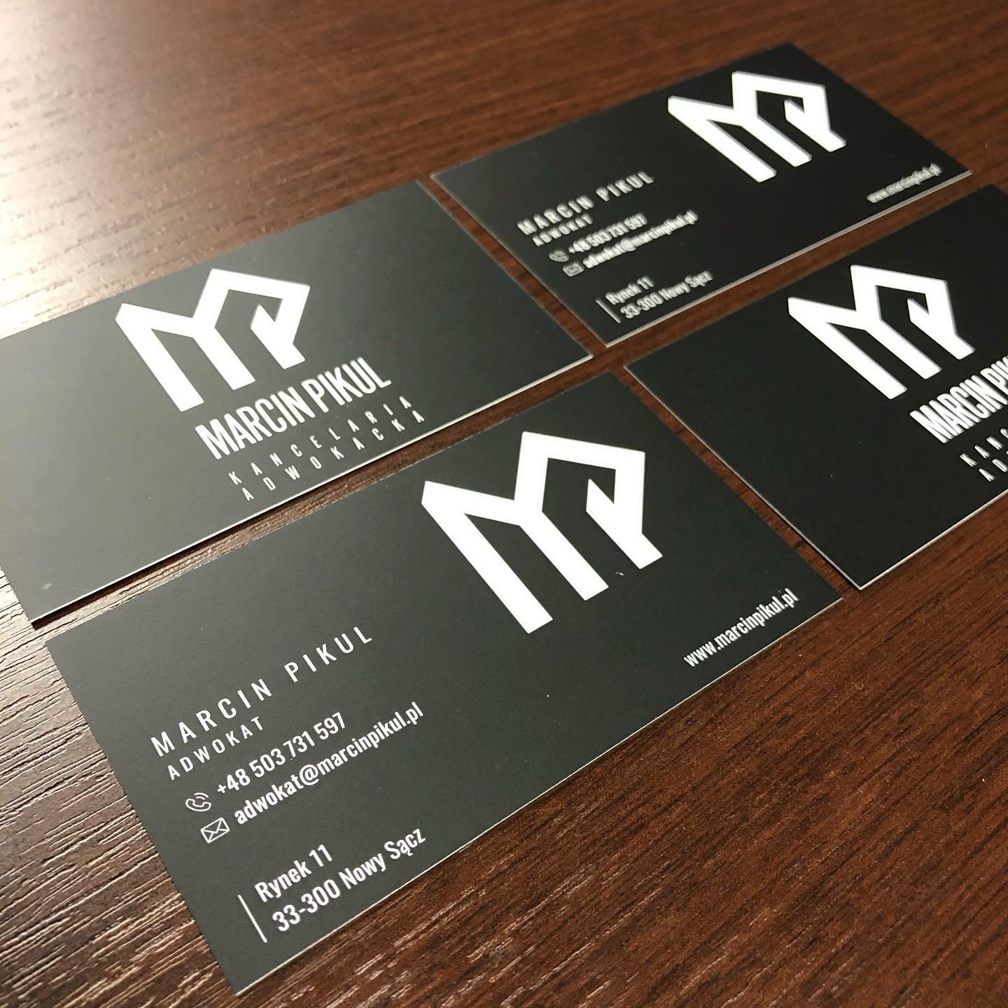 Cztery wizytówki adwokata Marcina Pikula leżące na drewnianym biurku, widoczne logo kancelarii w formie stylizowanej litery M.