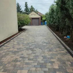 Ukończony podjazd z kostki brukowej w odcieniach szarości i beżu, z ciemnoszarym obramowaniem, prowadzący do garażu z brązową bramą, otoczony zielenią.