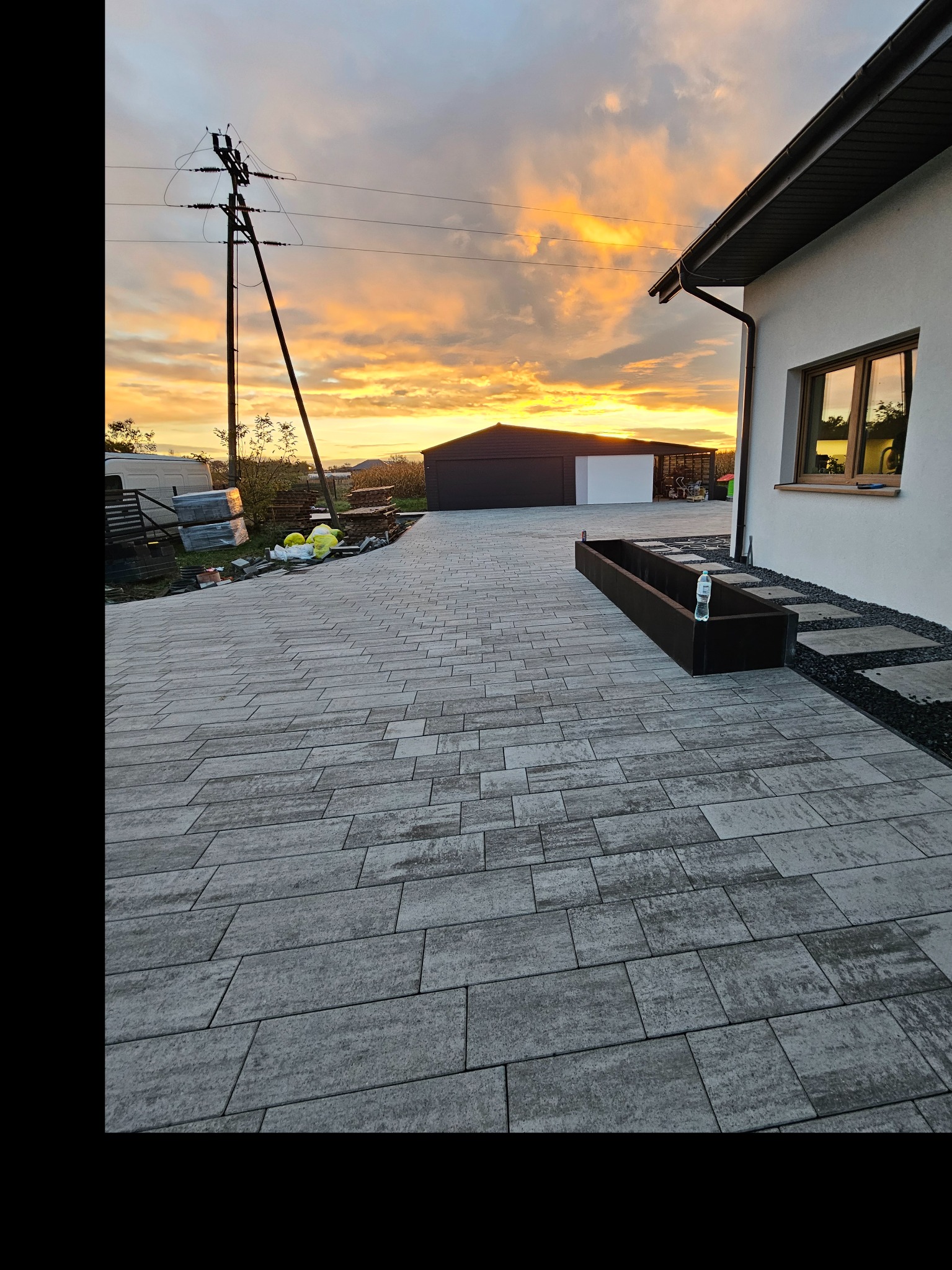 Ułożona kostka brukowa w odcieniach szarości prowadząca do garażu, z widocznym fragmentem domu i zachodem słońca w tle. Donica z butelką wody na kostce.