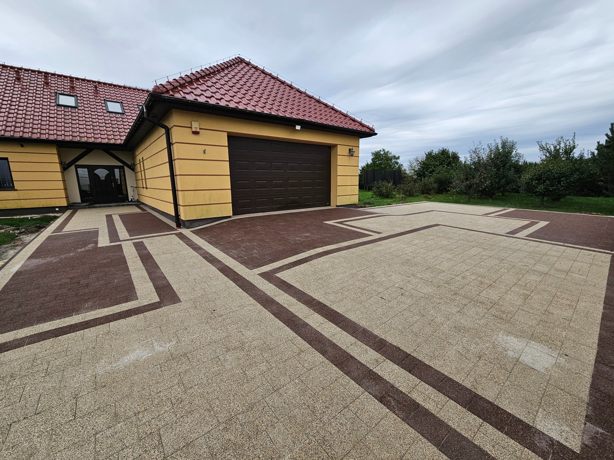 Dwukolorowa kostka brukowa ułożona w geometryczny wzór przed domem z garażem. Czerwona dachówka, beżowa elewacja. Pochmurne niebo w tle.
