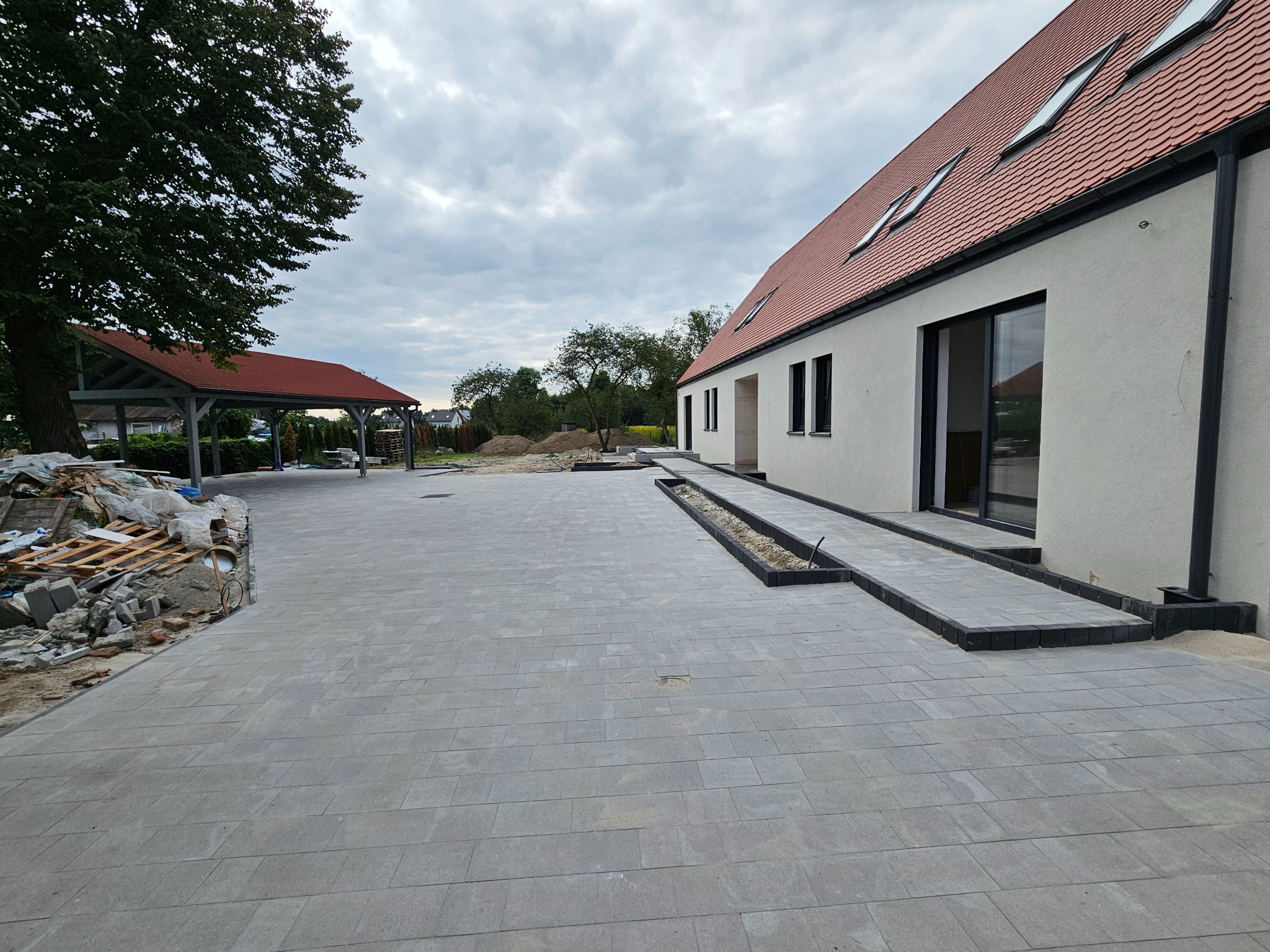 Ułożona szara kostka brukowa przed domem z czerwoną dachówką i wiatą. Widoczne pozostałości materiałów budowlanych. Ujęcie z poziomu gruntu.