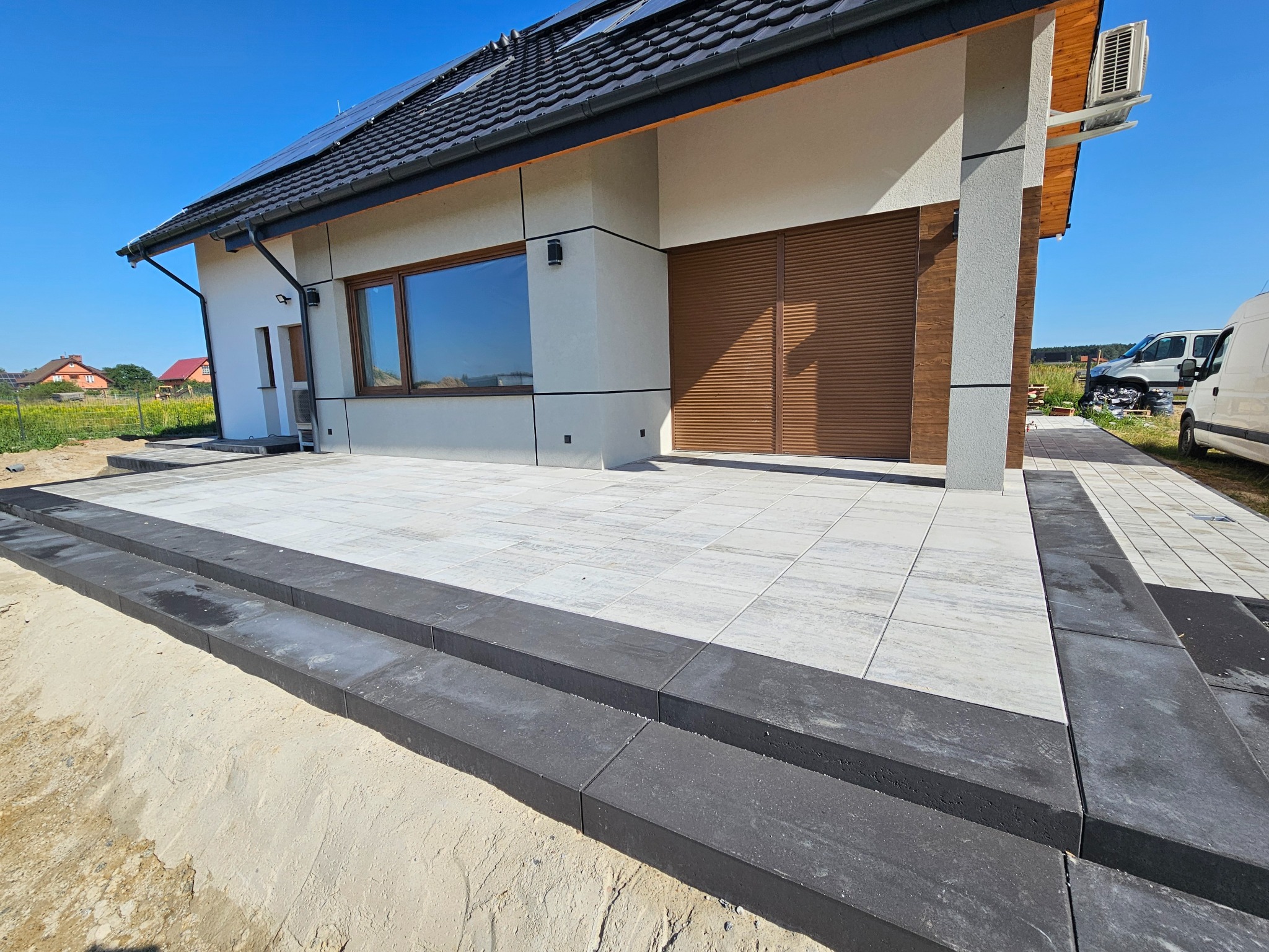 Nowoczesny taras z jasnoszarej kostki brukowej, obrzeża z ciemnoszarego kamienia, na tle domu z panelami słonecznymi i błękitnego nieba. Widok z niskiej perspektywy.