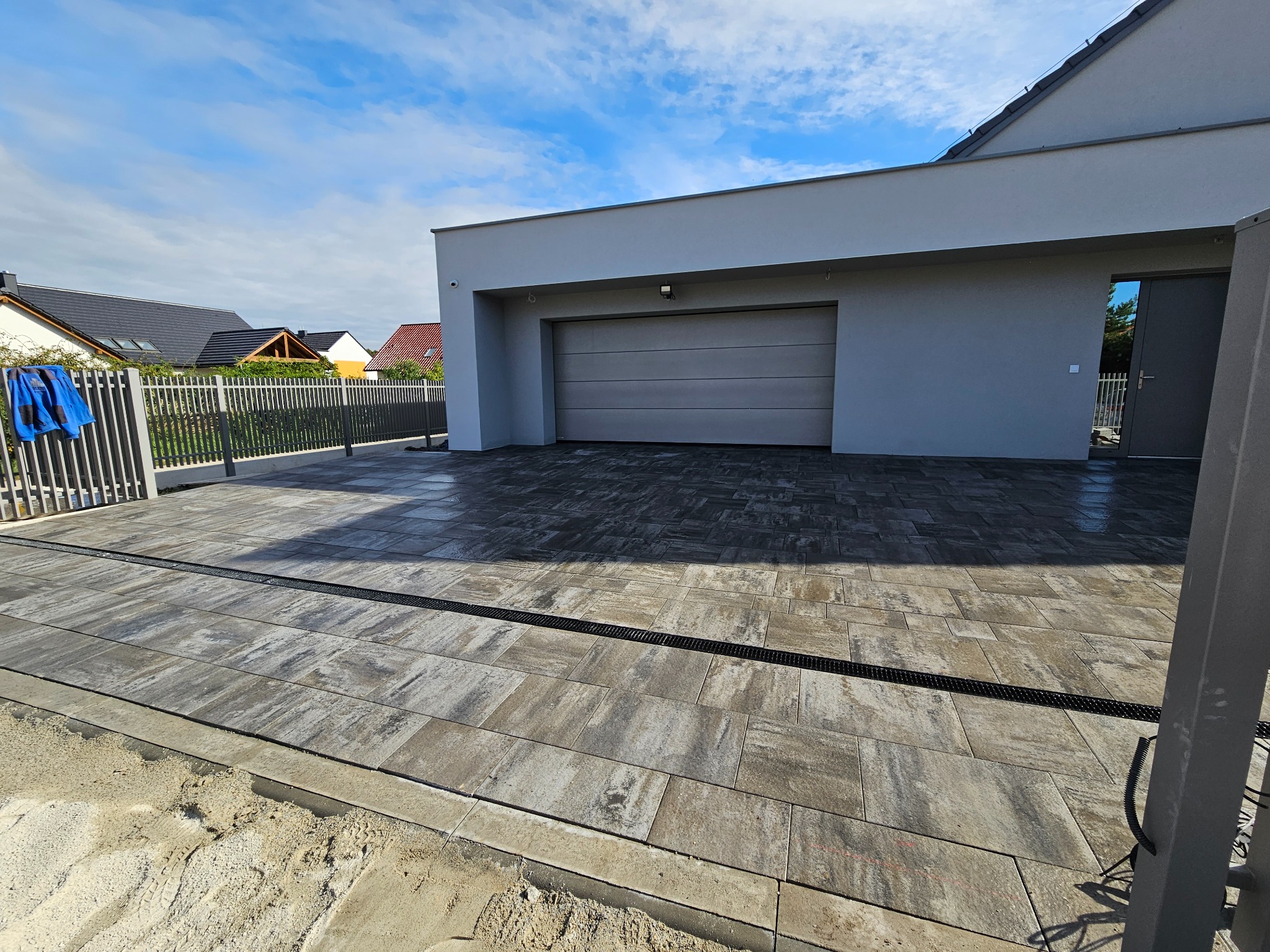 Nowoczesny podjazd z kostki brukowej w odcieniach szarości, z odpływem liniowym, prowadzący do garażu w domu jednorodzinnym. Widoczne prace wykończeniowe.