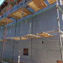 Budynek w trakcie ocieplania, ściany pokryte szarymi płytami izolacyjnymi, widoczne rusztowania i fragmenty ceglanej elewacji.