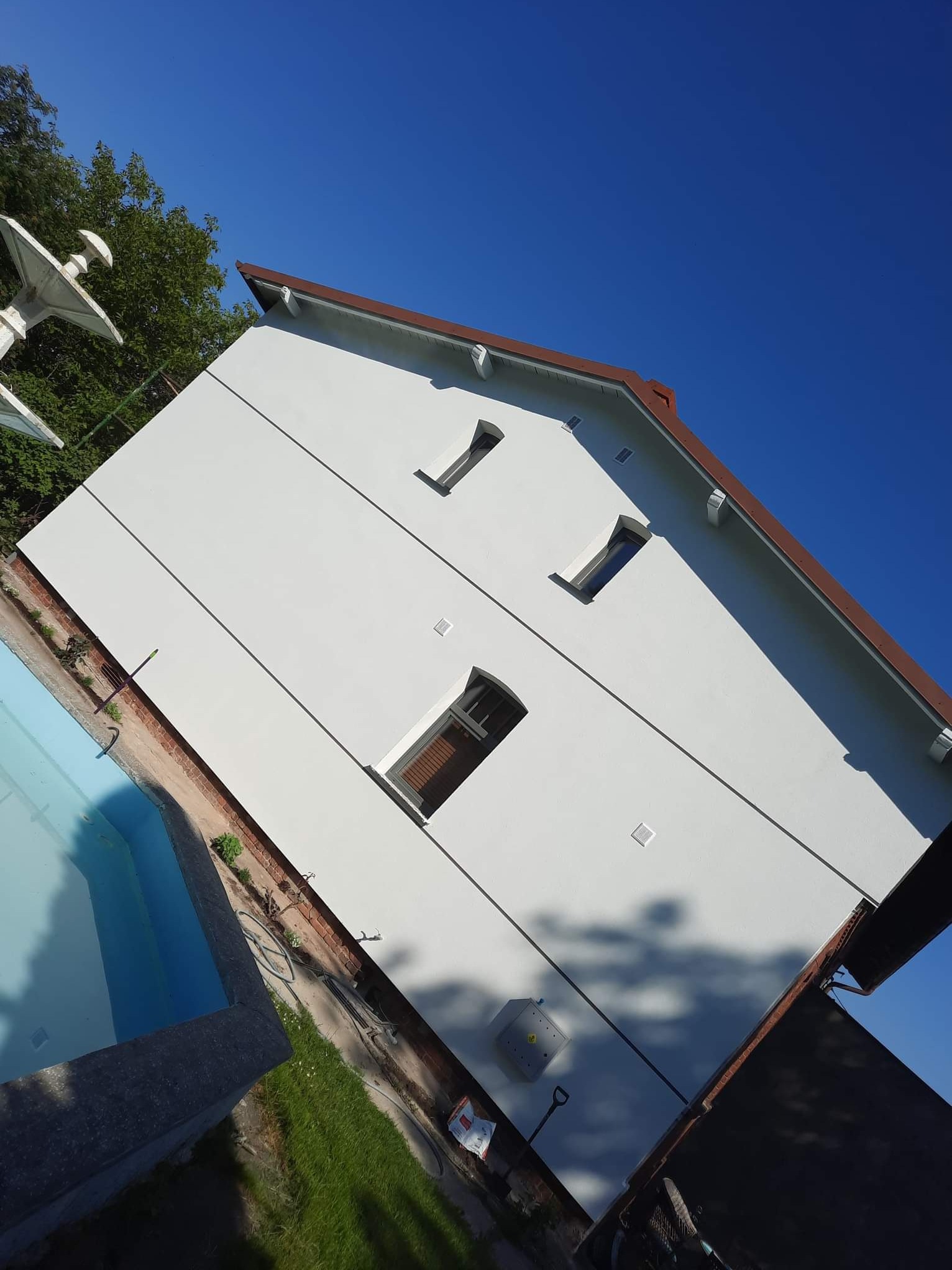 Boczny widok odnowionej, białej elewacji budynku z brązowym dachem, widzianej z poziomu basenu w słoneczny dzień. Widoczne trzy okna o różnej wielkości i kształcie.