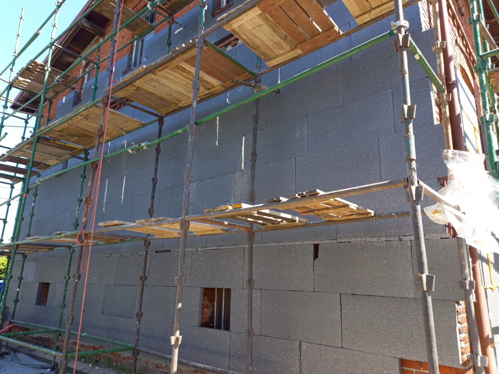 Budynek w trakcie ocieplania, ściany pokryte szarymi płytami izolacyjnymi, widoczne rusztowania i fragmenty ceglanej elewacji.