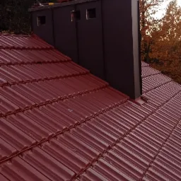 Czerwona dachówka ceramiczna na skośnym dachu z kominem wentylacyjnym w kolorze grafitowym, jesienne drzewa w tle.