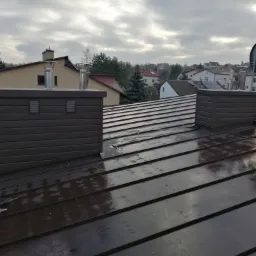 Nowy, błyszczący, ciemnobrązowy dach z blachy na tle pochmurnego nieba i zabudowań, widoczne dwa kominy wentylacyjne i narzędzia dekarskie.