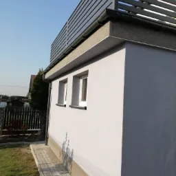 Nowoczesna bryła budynku z szarą elewacją, widoczne dwie białe okna, czarne rynny i metalowa konstrukcja na dachu, kostka brukowa wokół budynku.