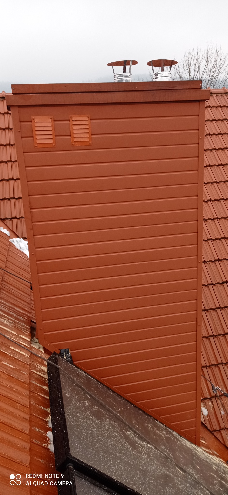 Nowe, brązowe obicie komina z wentylacją, dopasowane kolorystycznie do dachówki ceramicznej, widok z góry na tle pochmurnego nieba.