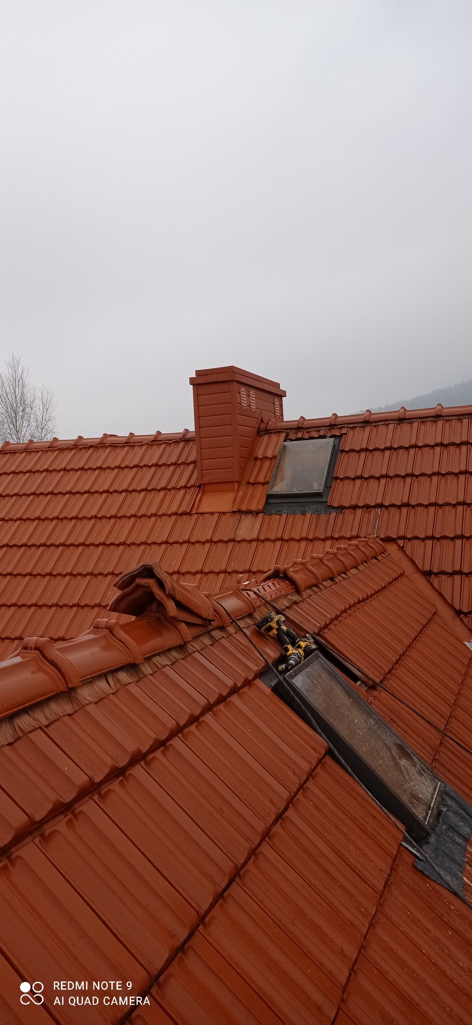 Czerwony dach ceramiczny z kominem i oknem dachowym w trakcie prac, widoczne narzędzia i fragmenty uszkodzonych dachówek, pochmurne niebo w tle.