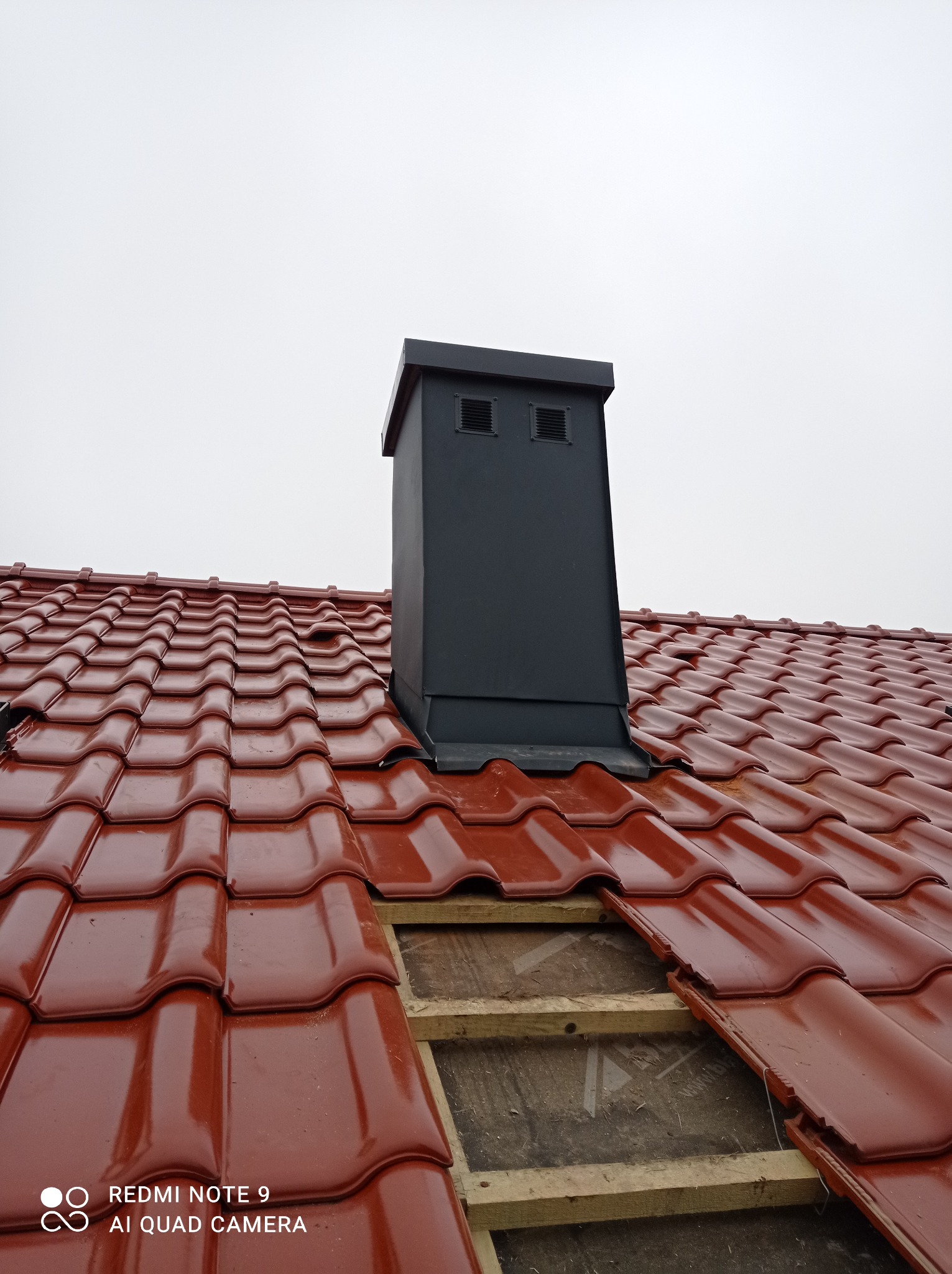 Czerwona dachówka ceramiczna z czarnym kominem wentylacyjnym, fragment dachu w trakcie wymiany z widoczną konstrukcją drewnianą.