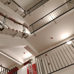 Moki Bud - Klatka schodowa w budynku, widok z dołu, białe ściany i schody, czarne metalowe balustrady, widoczne zabezpieczenia budowlane z czerwono-białych pasów.