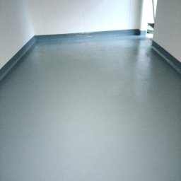 Moki Bud - Świeżo wylana, szara, samopoziomująca wylewka podłogowa w pomieszczeniu z białymi ścianami i szarymi listwami przypodłogowymi, widoczny fragment otwartych drzwi w tle.