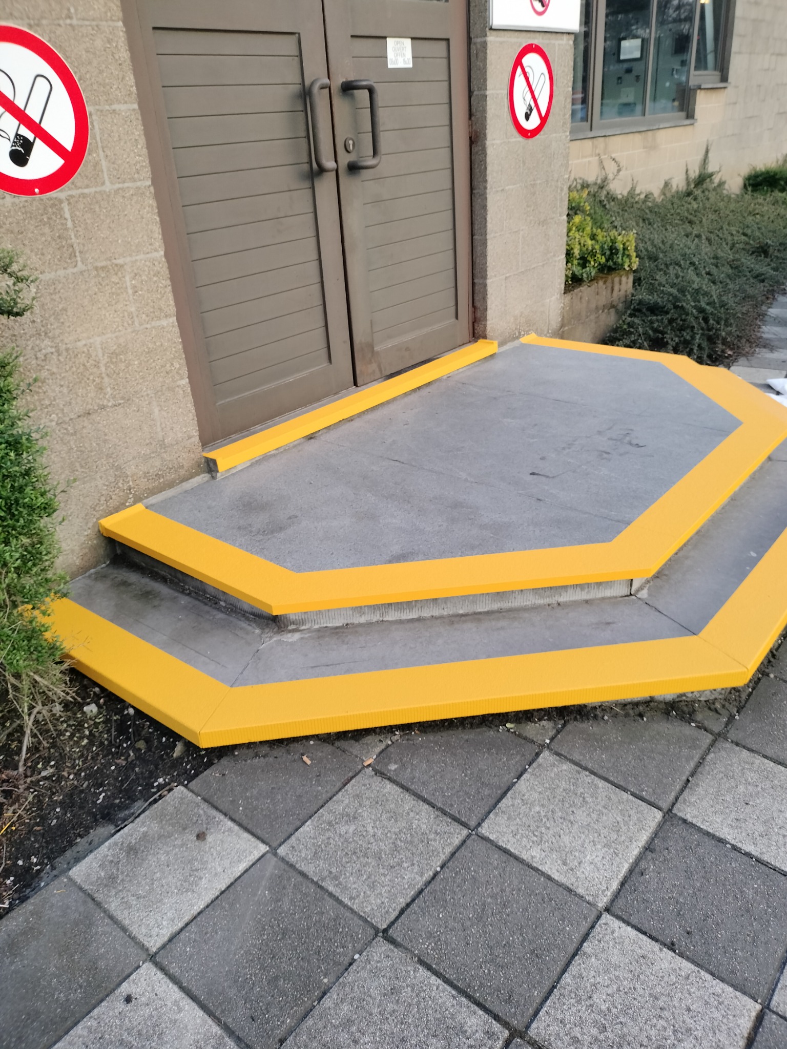 Zewnętrzne schody wejściowe z szarą nawierzchnią i jaskrawożółtymi krawędziami, przed wejściem do budynku z zakazem palenia.