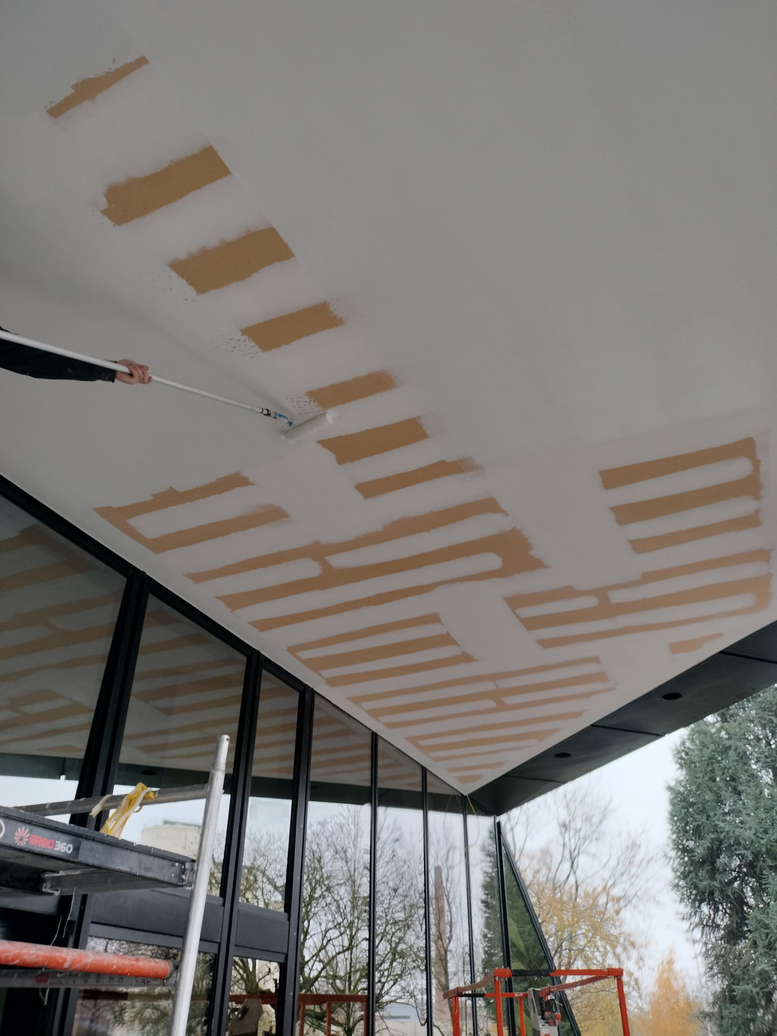 Malowanie sufitu w trakcie prac wykończeniowych, widoczne fragmenty surowego podłoża i świeżo nałożona biała farba, drabina i duże okna w tle.