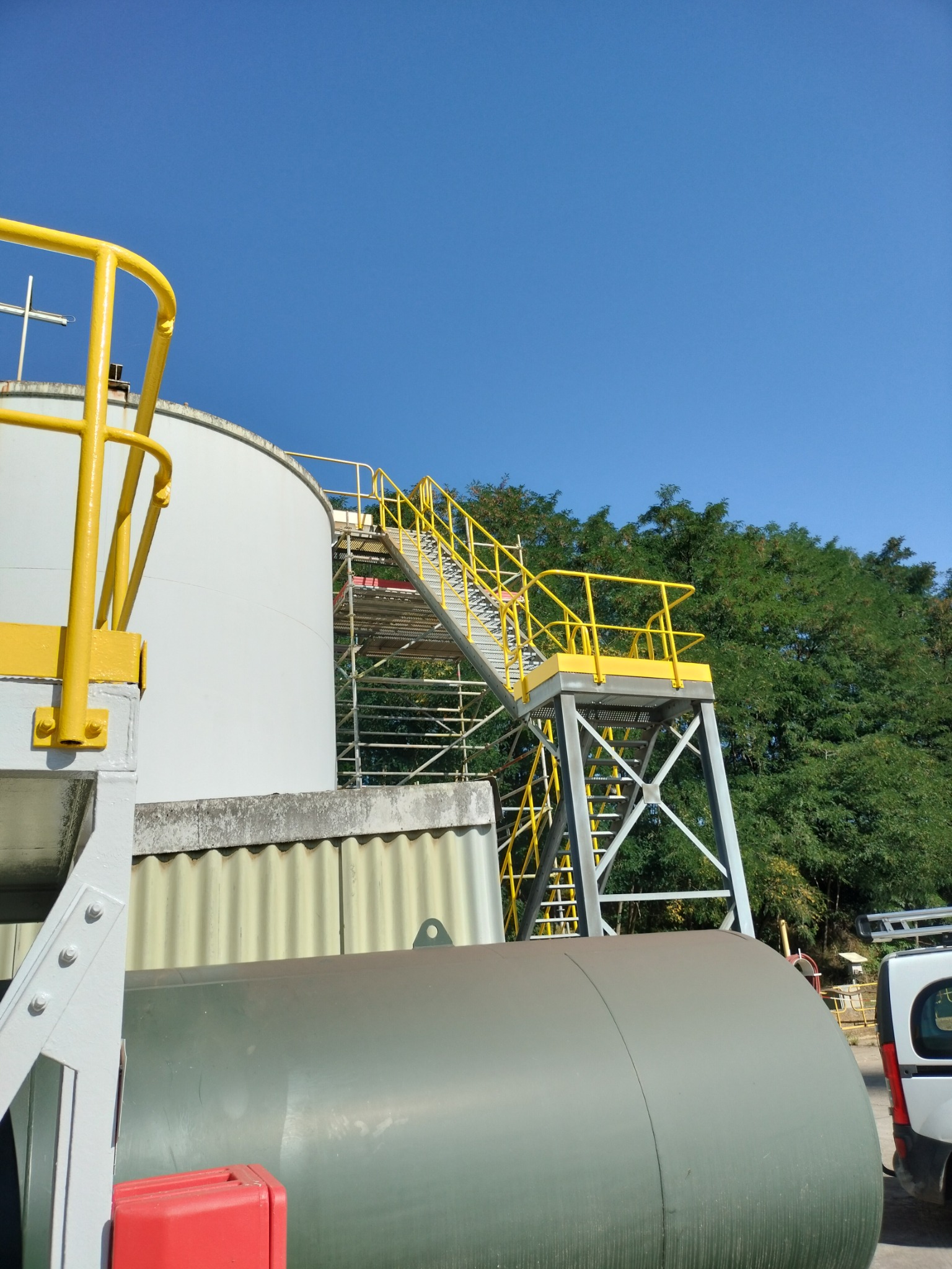 Zewnętrzny zbiornik przemysłowy z metalowymi schodami i żółtymi barierkami, na tle błękitnego nieba i zielonych drzew.